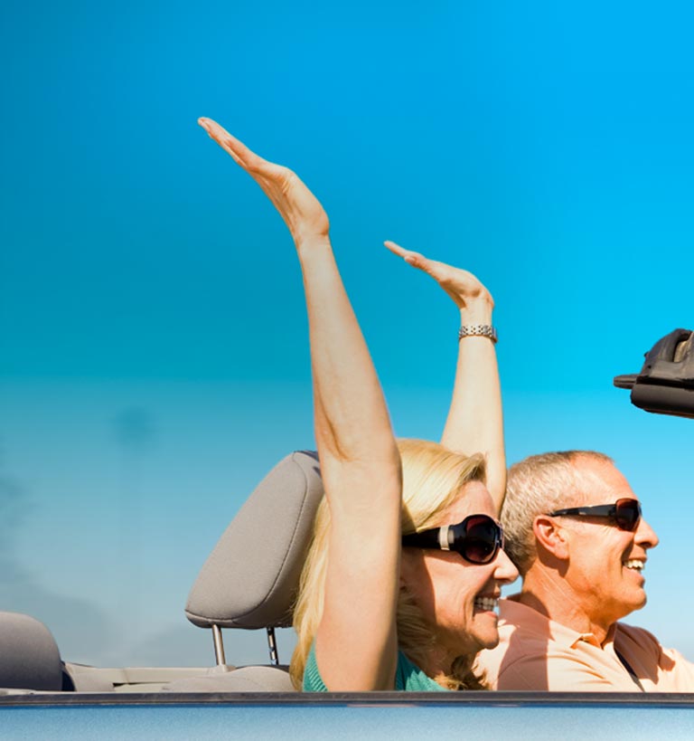 Happy older couple in convertible with hands raised.